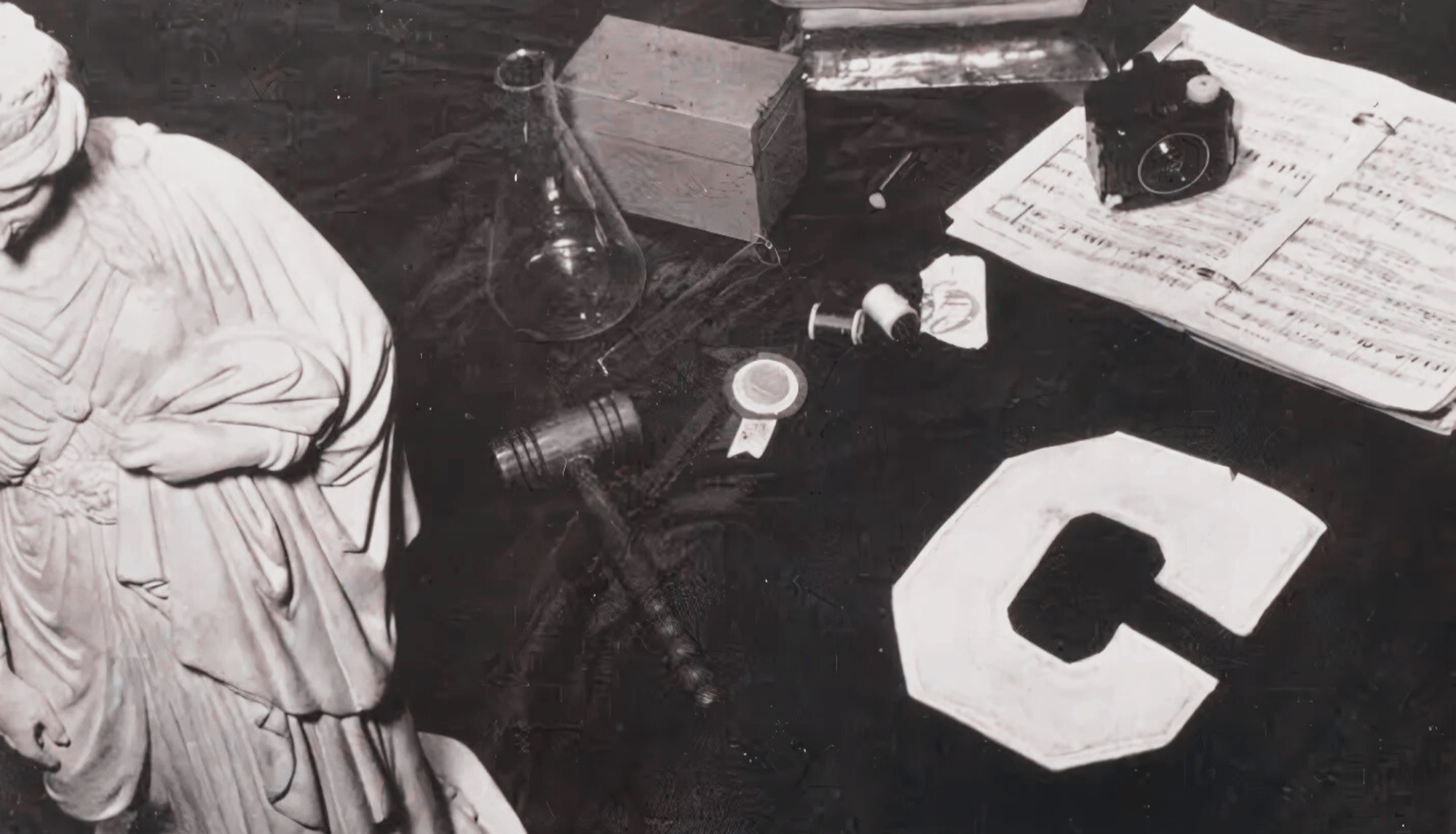 Black and white still life with classical statue, tools, and objects on a dark surface.