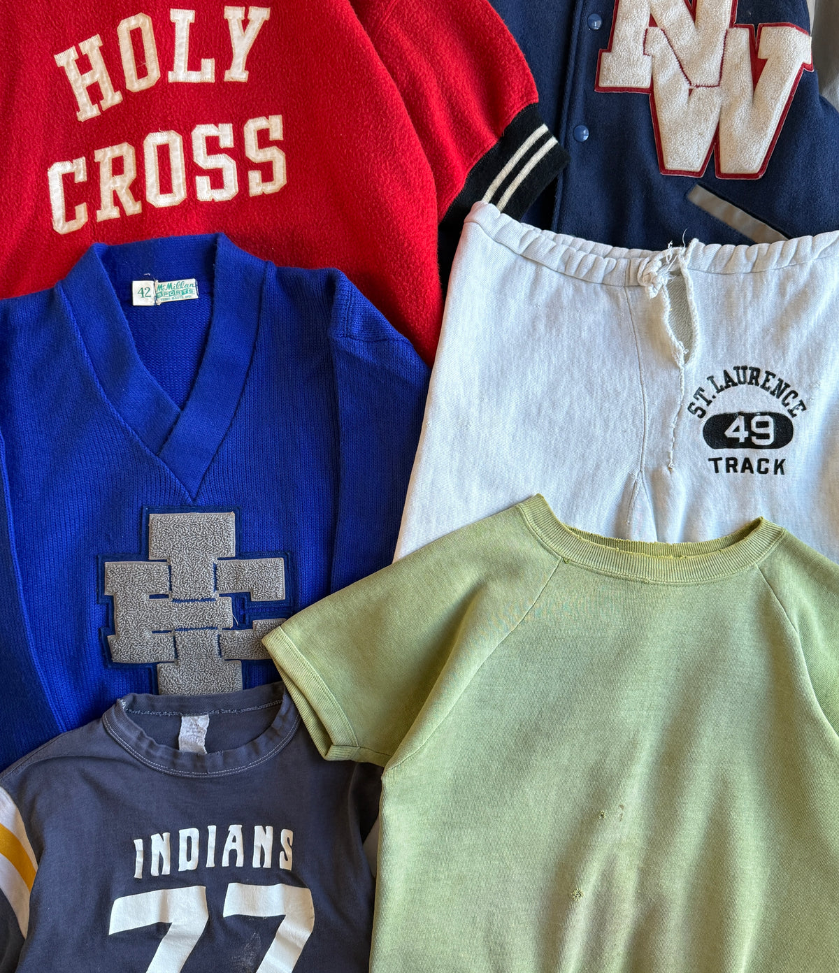 A flat-lay arrangement of six vintage athletic t-shirts and sweatshirts, featuring collegiate team designs including 'Holy Cross' in red, a blue V-neck with a gray 'UC' patch, a navy jacket with a 'W' on the chest, white track pants, a navy 'Indians 77' shirt, and a light green raglan t-shirt.